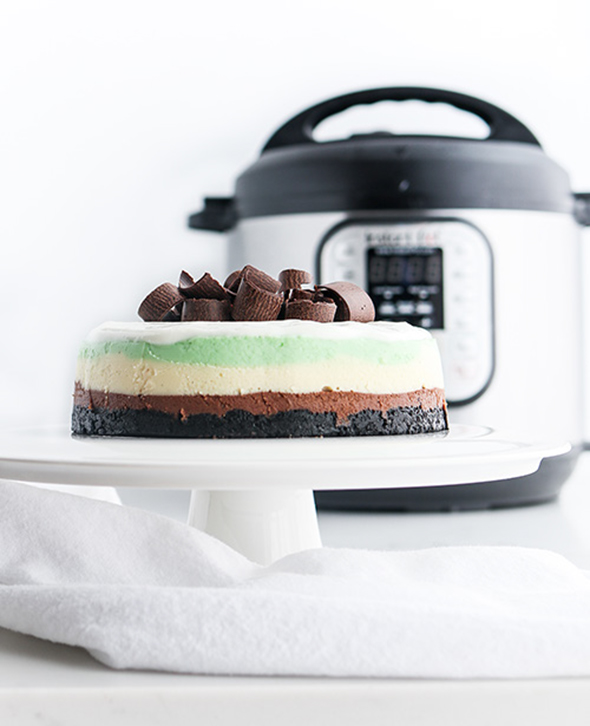 straight-on shot included in the Instantly Sweet Dessert Cookbook - featuring a white cake stand topped with triple layer mint cheesecake with chocolate curls and an Instant Pot in the background