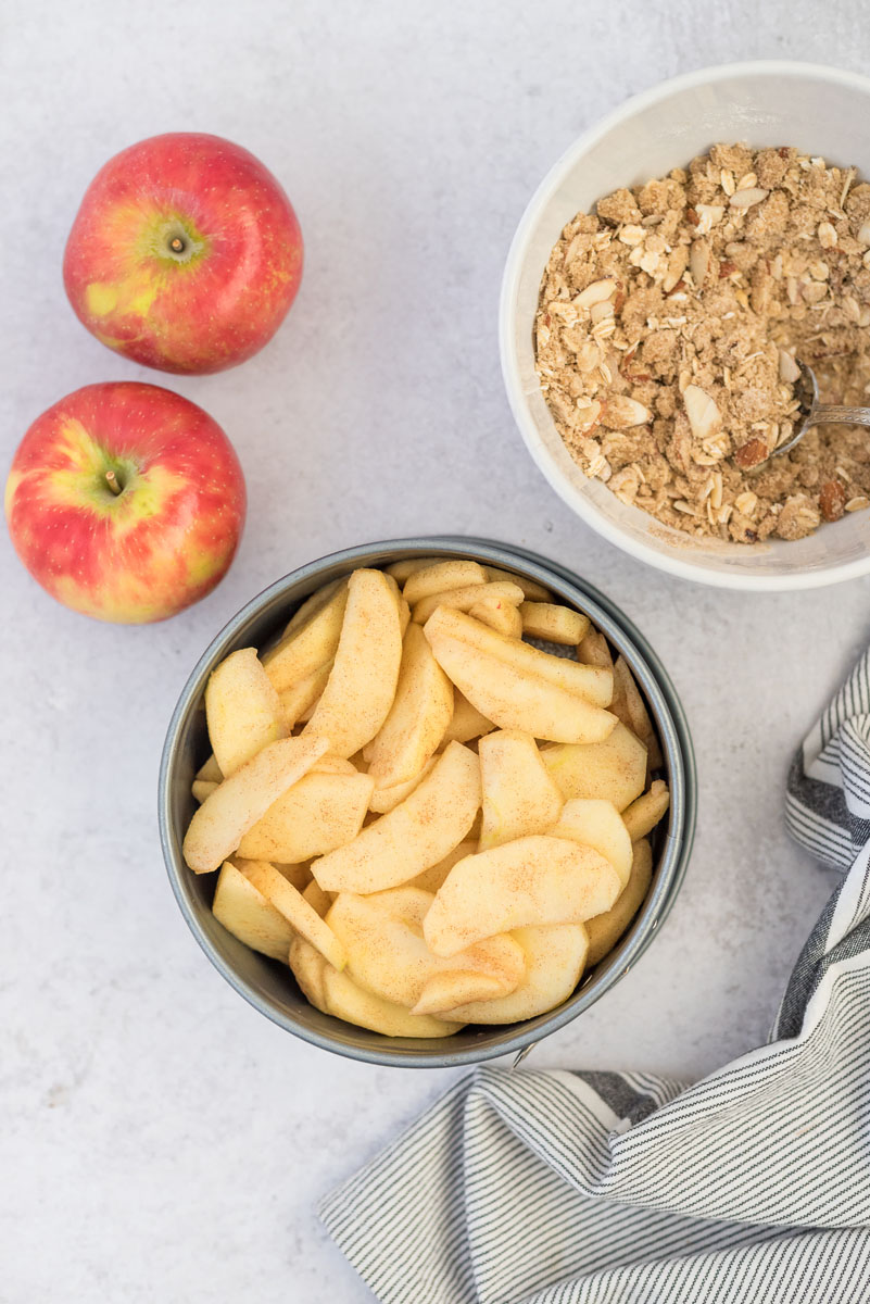 an overhead shot of diced apples in the springform pan with two red apples on the upper left a bowl full of the crumble topping on the right