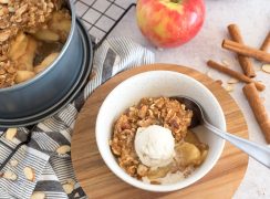 a shot of Instant Pot apple crisp in a white bowl, with a scoop of vanilla ice cream on top, with the pan of apple crisp, apples, and cinnamon sticks in the background