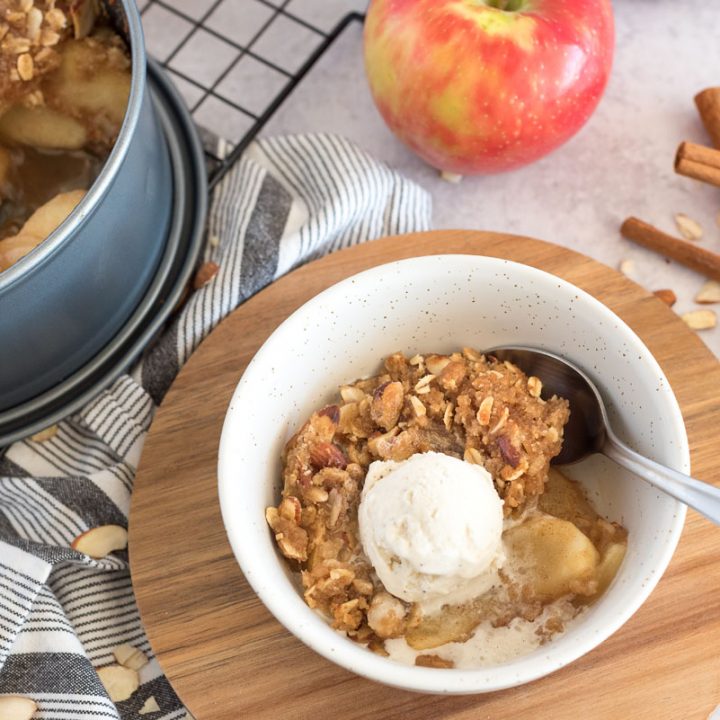 a shot of Instant Pot apple crisp in a white bowl, with a scoop of vanilla ice cream on top, with the pan of apple crisp, apples, and cinnamon sticks in the background