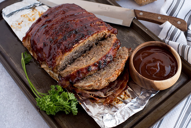 BBQ bacon Instant Pot meatloaf, sliced and ready to serve, pictured on a broiling pan with garnish, a knife, and extra BBQ sauce.