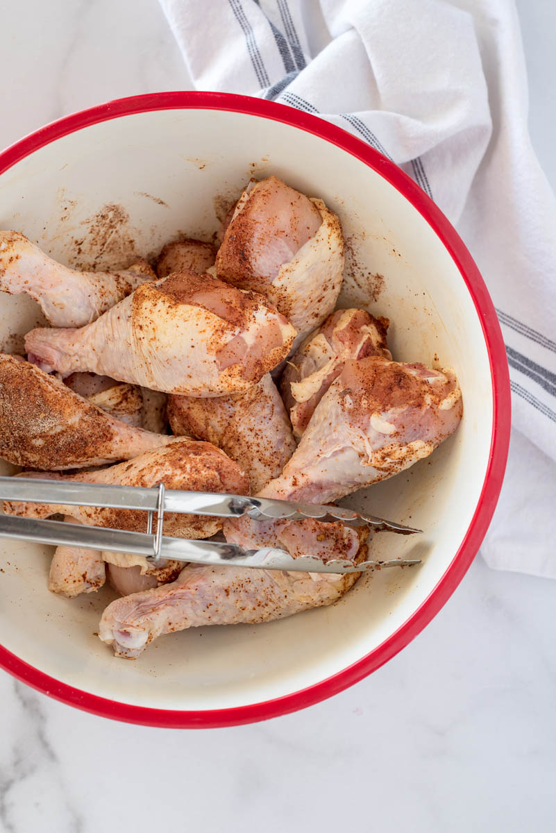 A white bowl with a red edge, containing uncooked chicken legs and seasoning against a white background