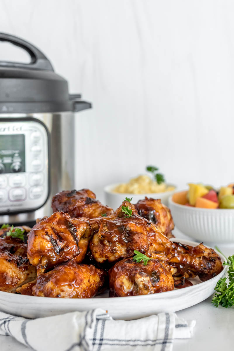 A photo of a white round plate of cooked BBQ chicken legs, with an Instant Pot and a bowl of fruit salad in the background