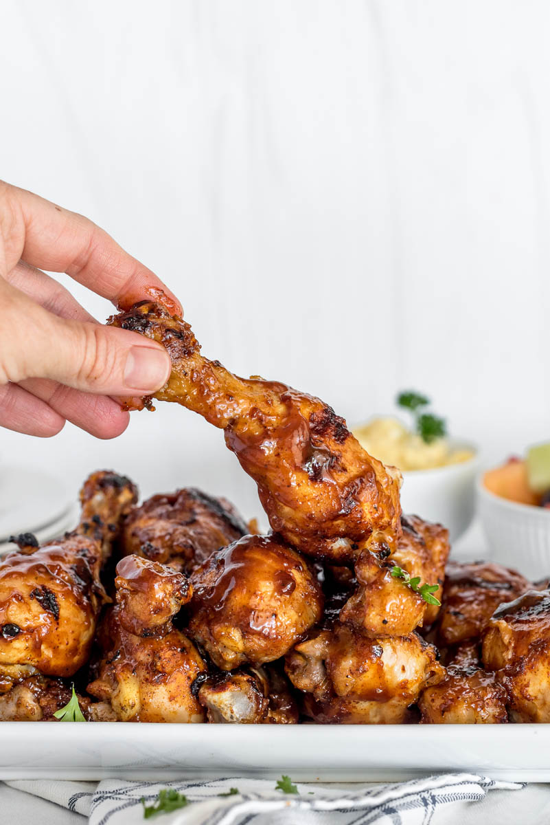 A hand picking up a chicken leg off of a white platter filled with lots of BBQ chicken legs made in the Instant Pot