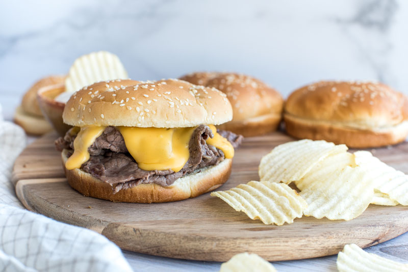 Instant pot shredded beef and melted cheddar cheese sandwiches on a wooden cutting board with ruffle cut potato chips