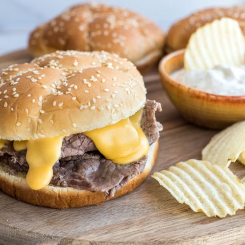 Instant pot beef and cheddar sandwiches on an onion roll with ruffle cut potato chips on a wooden cutting board