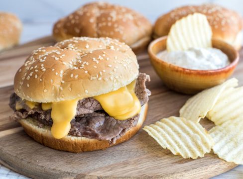 Instant pot beef and cheddar sandwiches on an onion roll with ruffle cut potato chips on a wooden cutting board