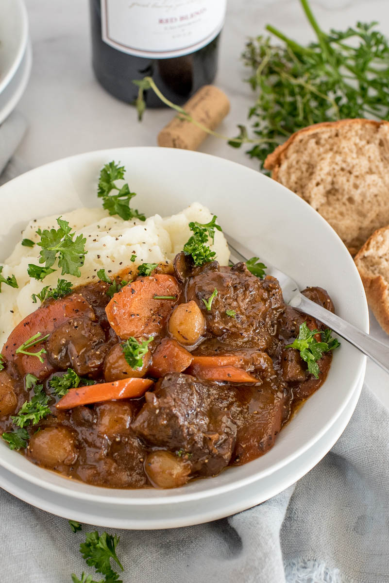 Close up picture of Instant Pot beef bourguignon cooked with carrots and pearl onions, served with mashed potatoes and fresh parsley. With rolls and a bottle of red wine in the background.