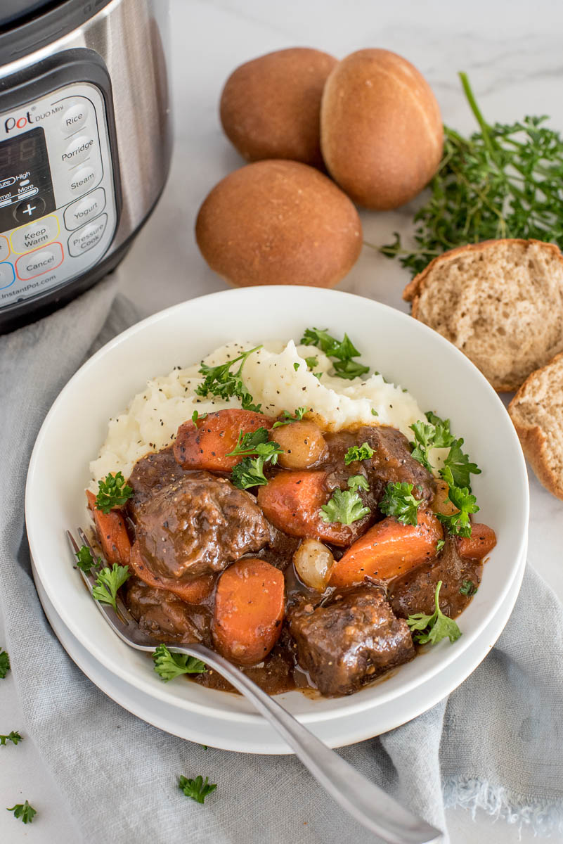 Overhead picture of beef bourguignon served in a white bowl with mashed potatoes and fresh parsley with an Instant Pot and rolls in the background.