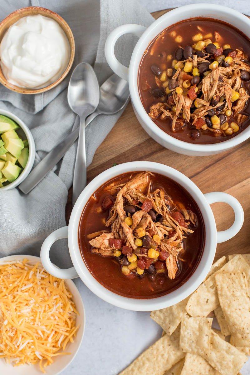 Overhead shot of Instant Pot chicken enchilada soup in a white bowl, with bowl of toppings including, cheese, avocado, and cilantro.