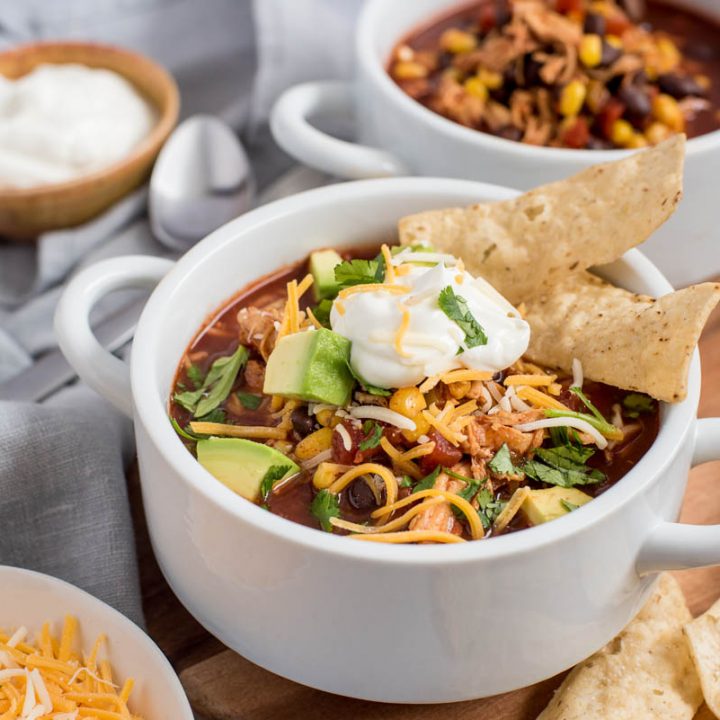 Close up shot of two bowls of Instant Pot chicken enchilada soup, one topped with cheese, sour cream, avocado, and chips, and the other without any toppings.