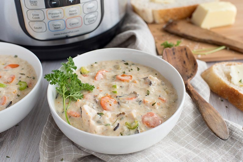 White bowl of creamy pressure cooker chicken wild rice soup garnished with parsley in front of an Instant Pot with white bread and a wooden spoon.