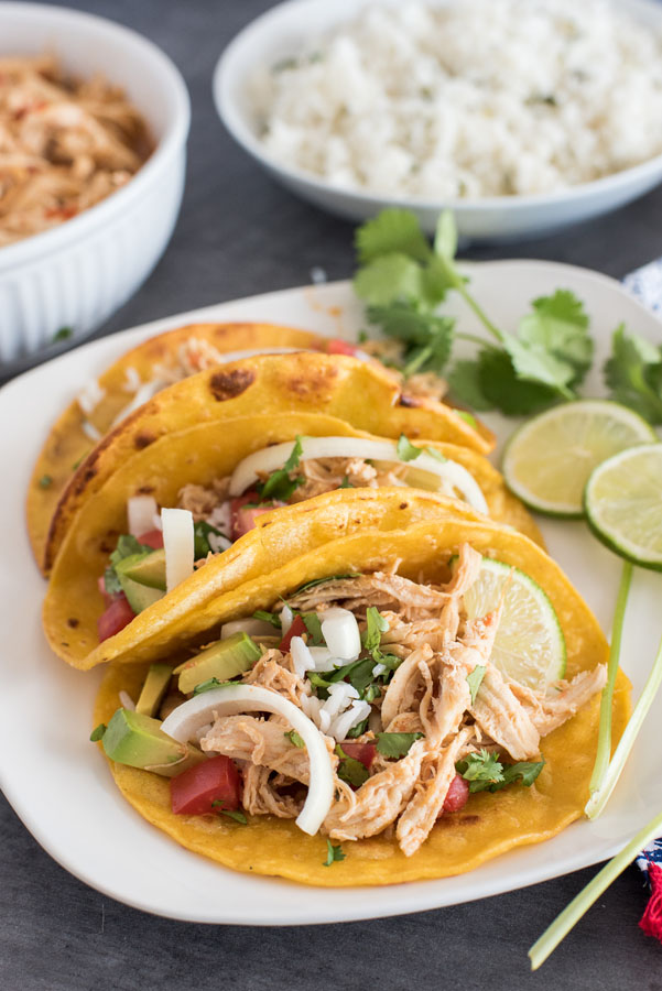 Three Instant Pot Tacos on a white plate, with a bowl of cilantro rice and shredded chicken taco filling in the background