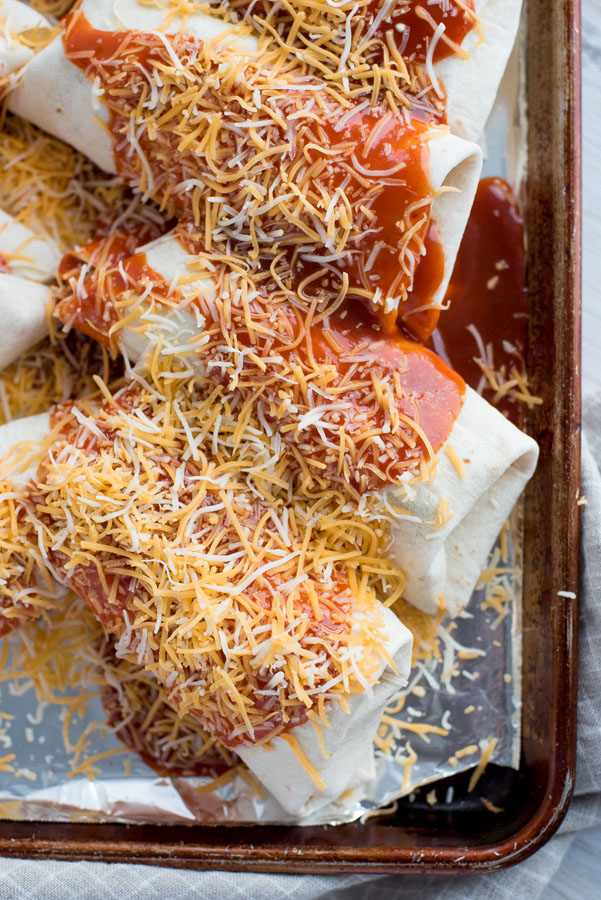 Close up on a baking sheet lined with tin foil holding Instant Pot Colorado Chili Burritos smothered with red enchilada sauce and shredded Colby jack cheese.
