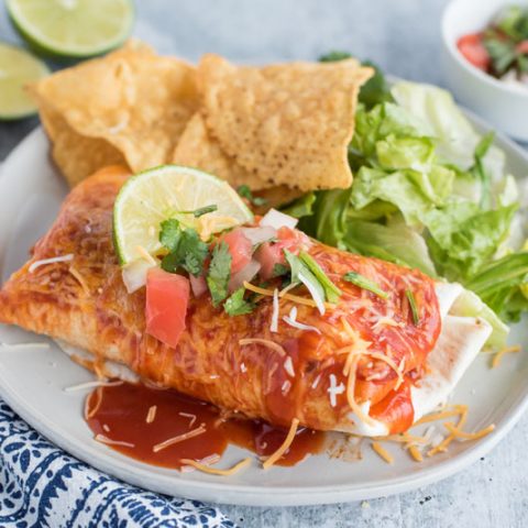 White dinner plate filled with tortilla chips, romaine lettuce and an Instant Pot Chili Colorado Smothered Burrito topped with melty cheese, lime wedge, fresh salsa and more enchilada sauce on a white background with a blue and white napkin and a silver fork.