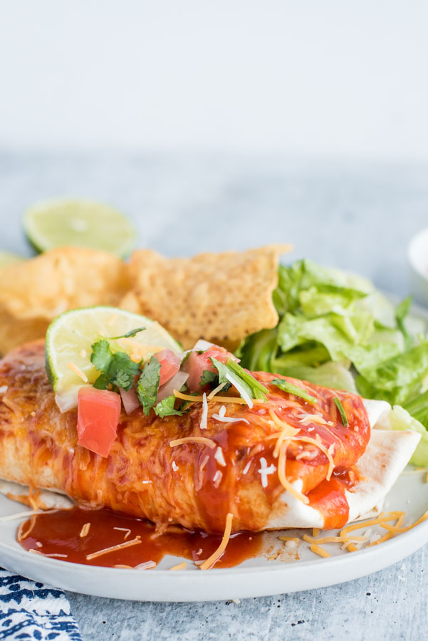 Close up on a freshly made Instant Pot / pressure cooker Chile Colorado smothered burrito with red echnilada sauce, melted Colby jack cheese, fresh salsa, cilantro, a lime wedge served with tortilla chips and crunchy romaine lettuce.