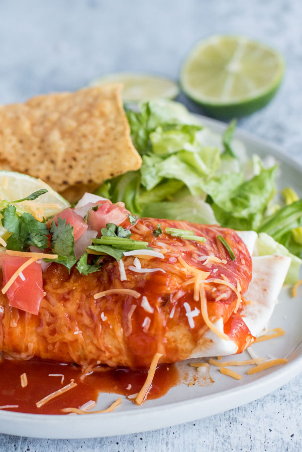 Close up on a white dinner plate with a freshly made Instant Pot / Pressure Cooker Chile Colorado Burrito smothered in red enchilada sauce, melted Colby jack cheese, fresh salsa and lime, served with tortilla chips and romaine lettuce.