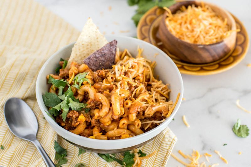 A close-up 45 degree shot of a white bowl of pressure cooker chili pasta, sitting on a yellow napkin garnished with cilantro, with a small bowl of grated yellow cheese visible in the background