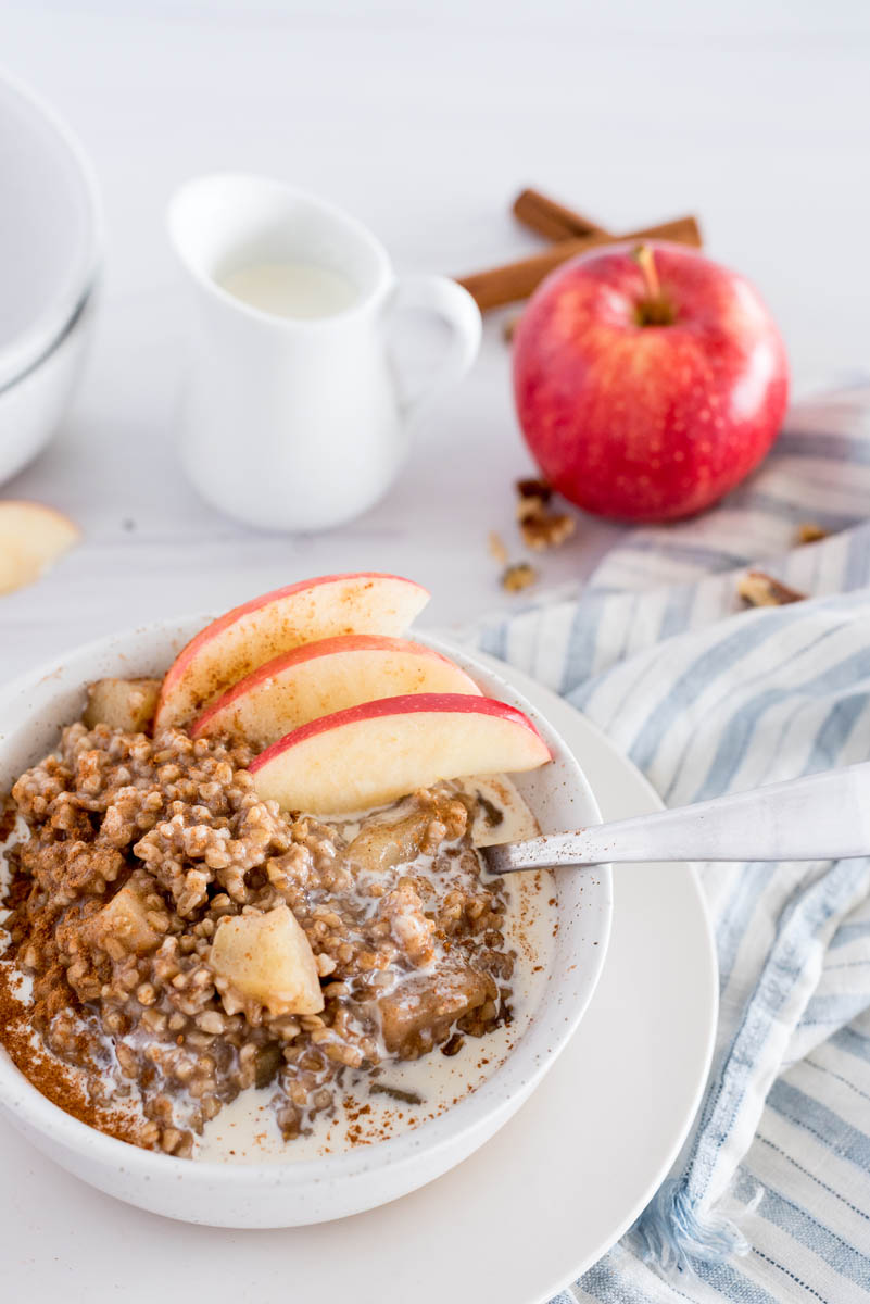 Close up picture of Instant Pot Cinnamon Apple Steel Cut Oats garnished with fresh sliced apples, in a white bowl, with a whole apple and a small pitcher of milk in the background. 