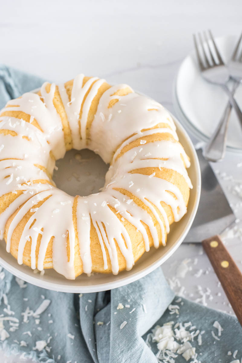 An overhead shot of coconut bundt cake, with a simple white coconut glaze sitting on a rimmed cake pan, with a blue napkin, serving spatula, white plates, and forks in the background