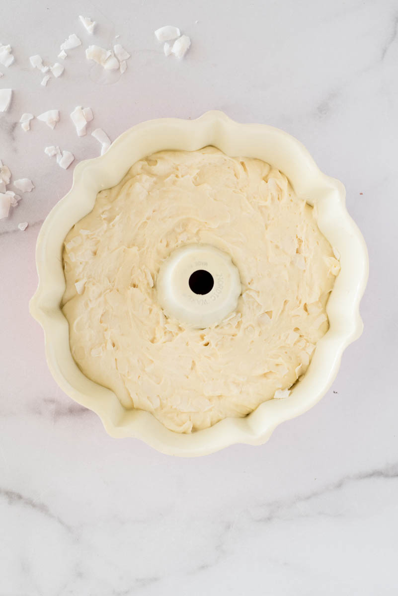 overhead shot of a cream-colored bundt pan filled 3/4 of the way with cake batter, with some coconut flakes spread on the top left corner. 