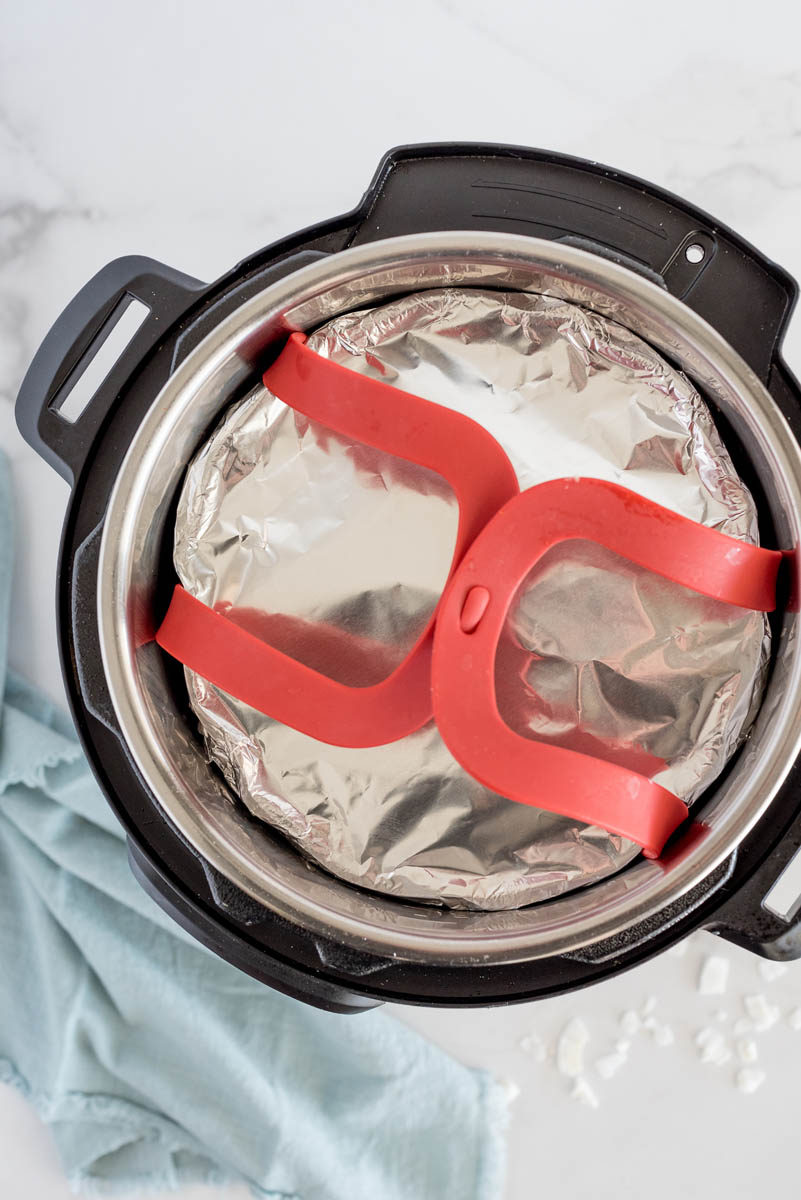 overhead shot of the Coconut Bundt Cake wrapped in foil and lowered with a sling into the Instant Pot, with a blue napkin in the lower left