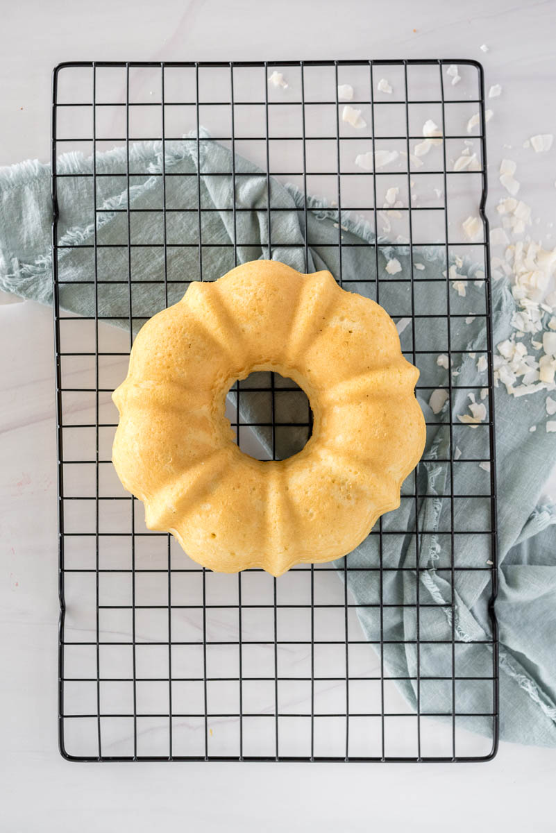 The cooked Coconut Bundt Cake set on a wire cooling rack with a blue napkin and coconut flakes in the background