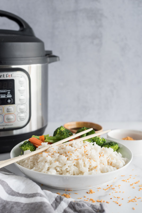 Side view of a white bowl of instant pot coconut rice topped with toasted coconut flakes next to steamed broccoli and carrots on a white and grey background in front of an instant pot pressure cooker