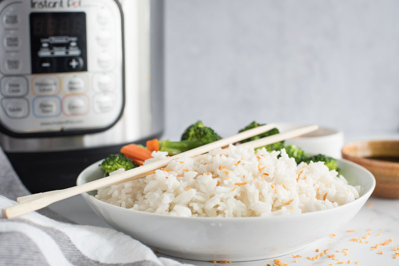 Side view of a white dish filled with instant Pot coconut rice topped with toasted coconut flakes and wooden chopsticks in front of an electric pressure cooker.