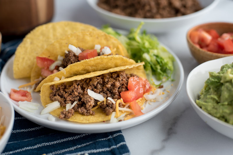 Instant Pot tacos in hard shells on a white plate with tomatoes, onions, cheese, and lettuce, next to bowls with guacamole and tomatoes.