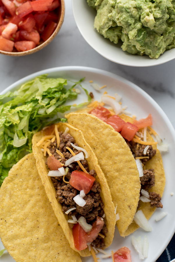 Overhead shot of Instant Pot ground beef tacos in a hard shell on a white plate with lettuce and tomatoes with a bowl of guacamole and tomatoes.