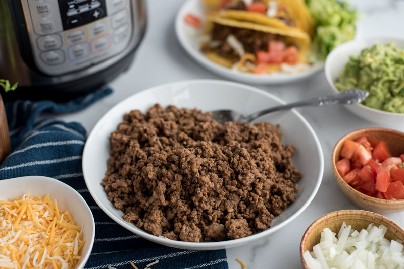 45 degree angle of ground beef in a bowl surrounded by bowls of guacamole, tomatoes, onions, and shredded cheese placed in front of an Instant Pot.