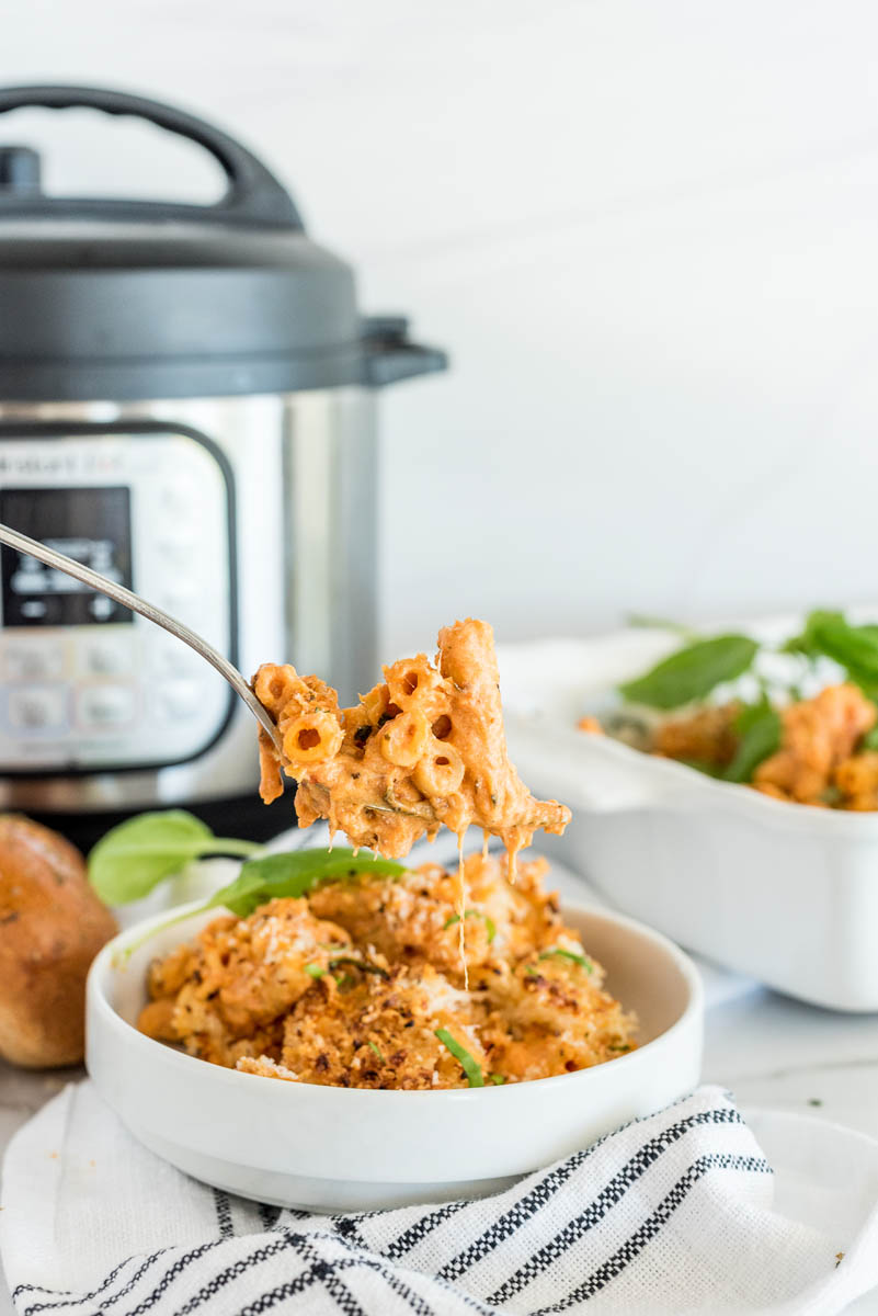 A direct shot of a white bowl of baked ziti with a spoonful of pasta being scooped out, in front of an Instant Pot