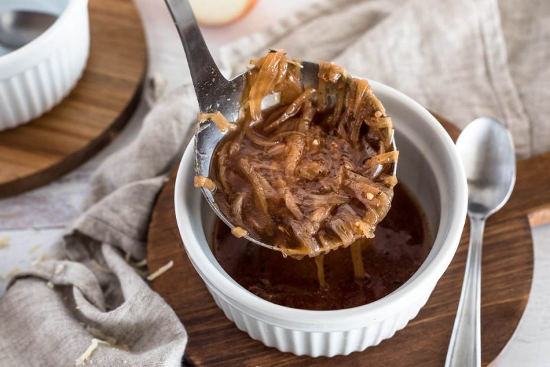 A silver ladle pouring rich brown French onion soup into a white ramekin, set on top of a wooden paddle with a silver spoon and gray napkin in the background