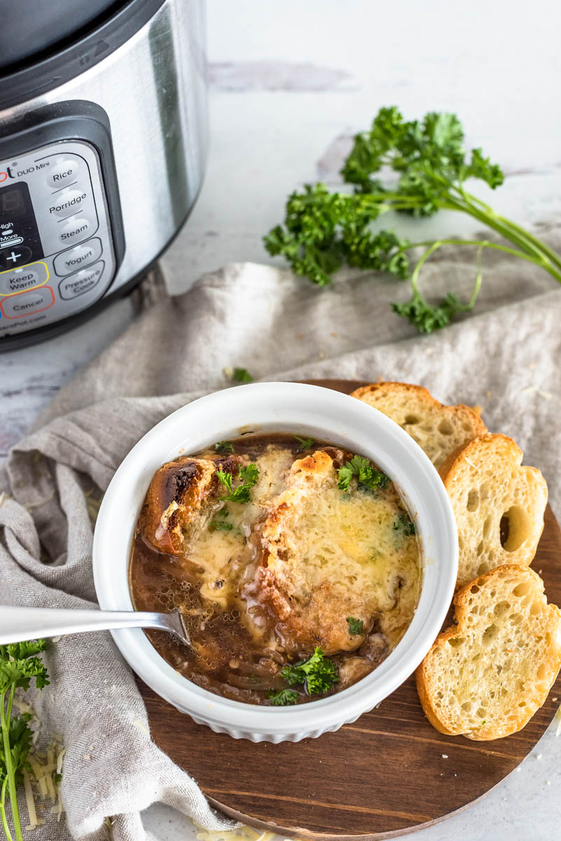 An overhead shot of pressure cooker French Onion Soup dished up in a white ramekin with lots of browned bread and cheese on top, with additional slices of bread, parsley, and an INstant Pot in the background