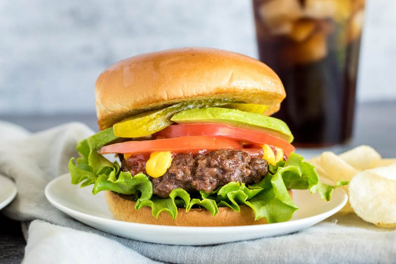 A thick, stacked hamburger, with the patty cooked in the Instant Pot, topped with lettuce, tomato, mustard, pickles, avocado, and a toasted bun on a white plate with chips and a tall glass of cola in the background