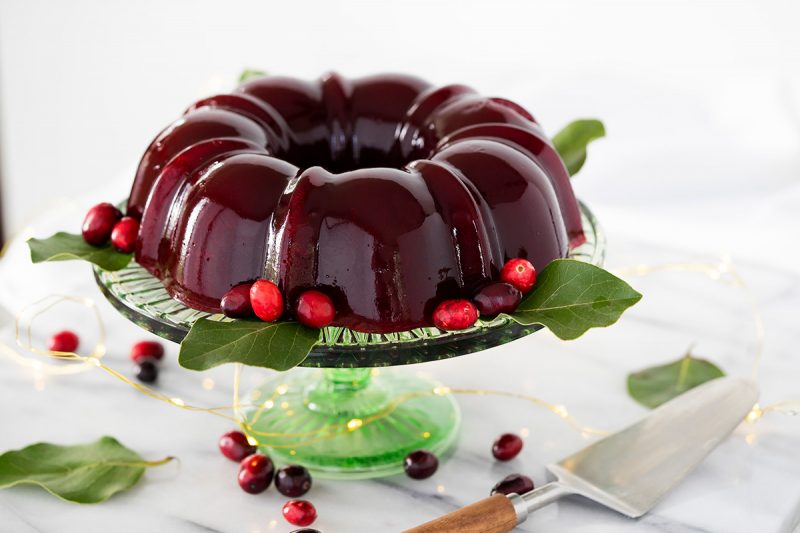 Deep red homemade cranberry jelly in the shape of a bundt pan on top of a green glass cake stand, garnished with bright red cranberries and leaves