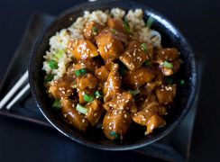 A horizontal close-up of Honey Sesame Chicken in a dark black bowl against a dark black background, with chopsticks tucked under the bowl