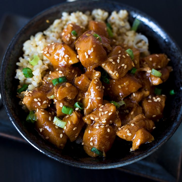 A horizontal close-up of Honey Sesame Chicken in a dark black bowl against a dark black background, with chopsticks tucked under the bowl