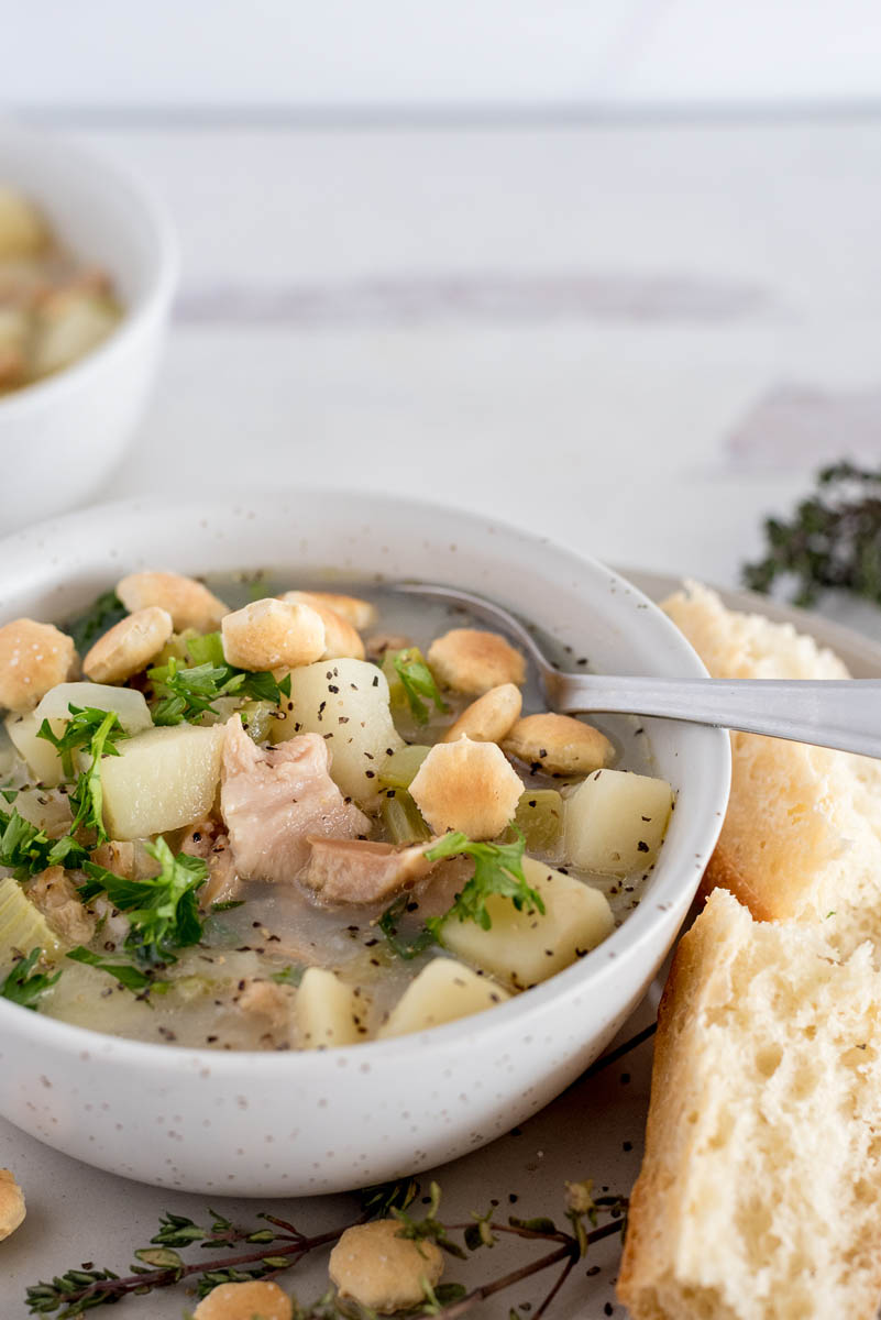 Close up of a white bowl with Instapot clam chowder, garnished with oyster crackers and fresh parsley, and served with a roll.