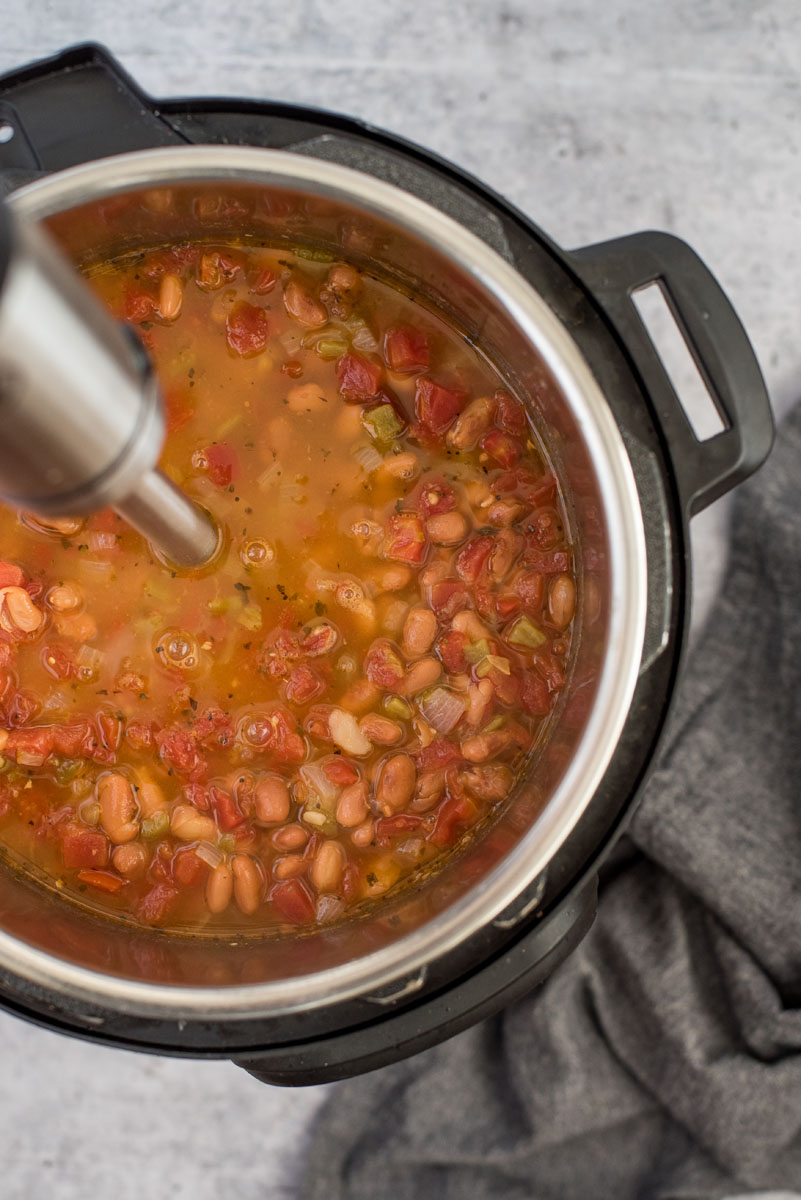 overhead shot of Instant Pot Pinto Bean Soup, with an immersion blender blending someo fo the beans, bacon, and tomatoes in the broth