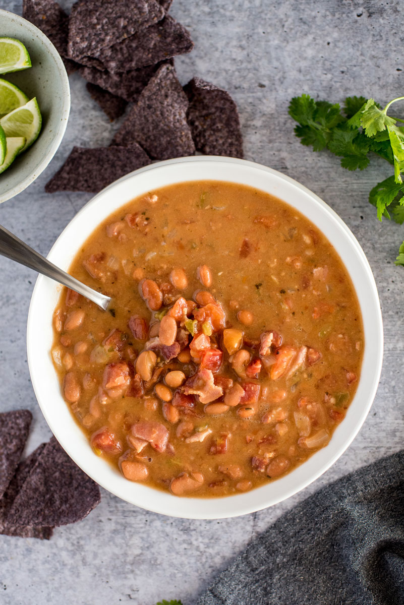 overhead vertical shot of Mexican Pinto Bean Soup, featuring a white dish filled with chunky charro bean soup topped with bacon and chopped cilantro on a stone gray backgrouond, with blue corn tortilla chips, limes, and cilantro in the background.