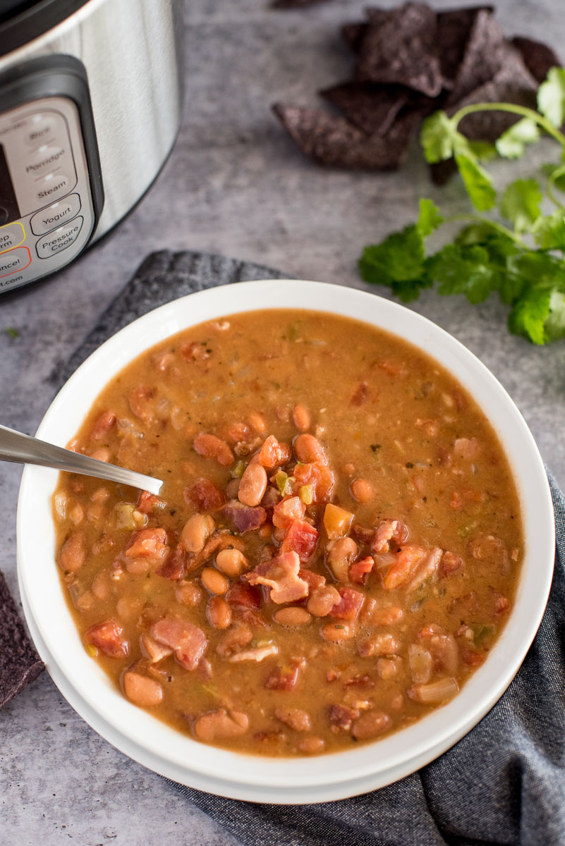 overhead shot of Instant Pot Mexican Pinto Bean soup, featuring a white bowl filled with chunky charro bean soup, topped with bacon and chopped cilantro on a stone gray backgrouond, with blue corn tortilla chips, cilantro, and an Instant Pot in the background.