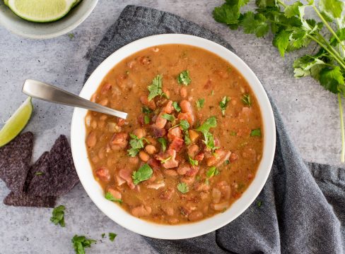 overhead shot of a white dish filled with chunky charro bean soup, topped with bacon and chopped cilantro on a stone gray backgrouond, with blue corn tortilla chips, limes, and cilantro in the background.