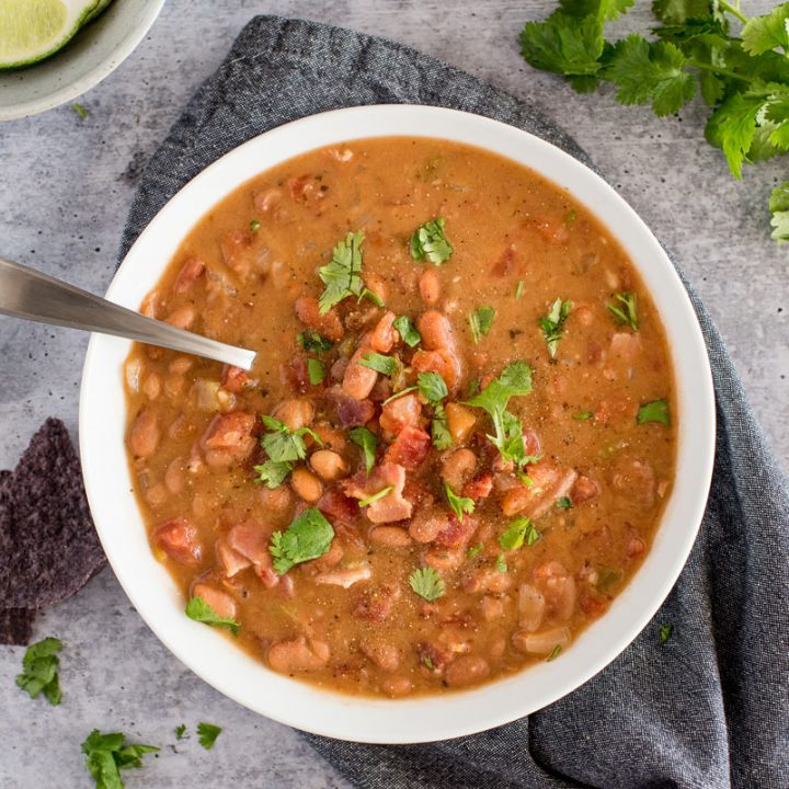 overhead shot of a white dish filled with chunky charro bean soup, topped with bacon and chopped cilantro on a stone gray backgrouond, with blue corn tortilla chips, limes, and cilantro in the background.