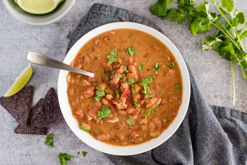 overhead shot of a white dish filled with chunky charro bean soup, topped with bacon and chopped cilantro on a stone gray backgrouond, with blue corn tortilla chips, limes, and cilantro in the background.