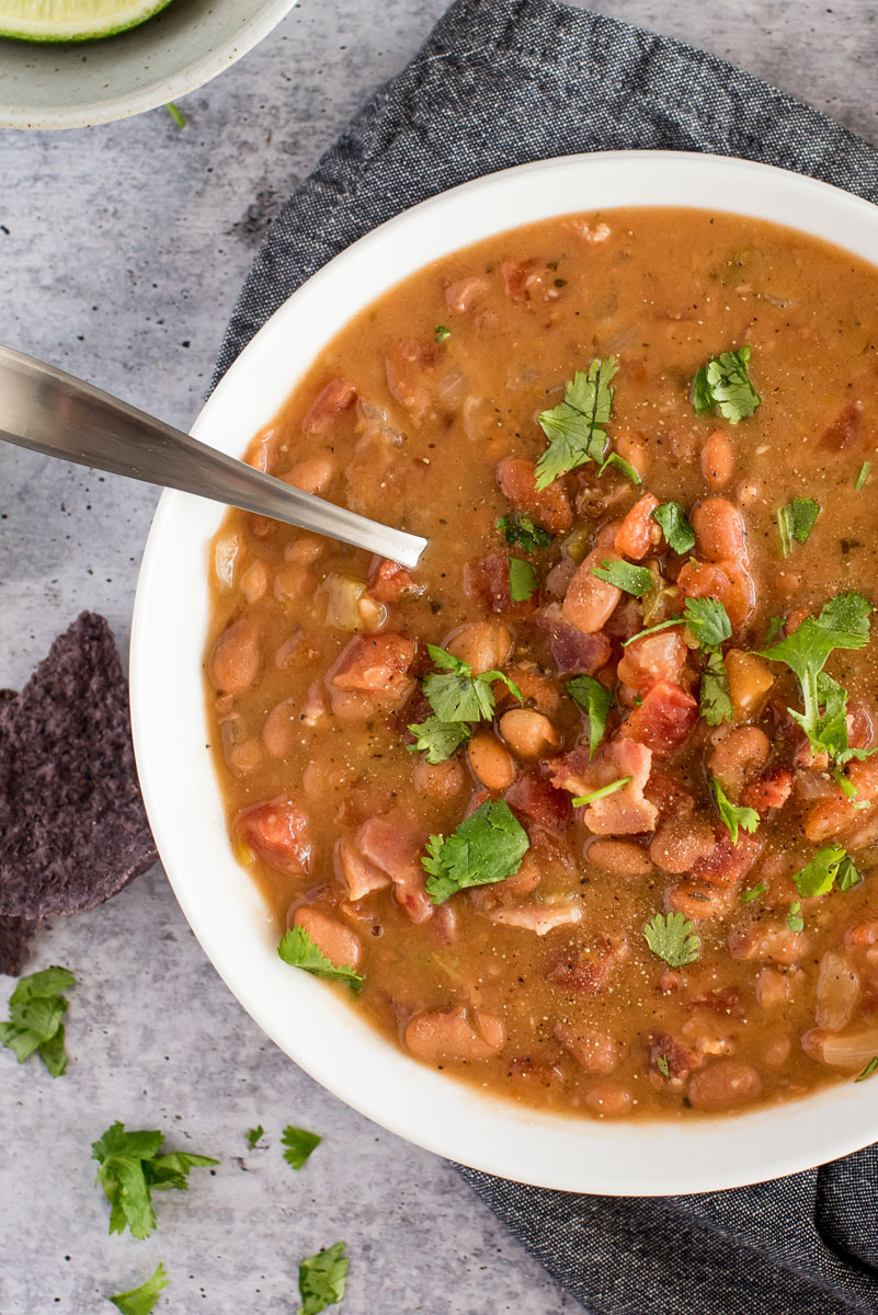 close-up overhead shot of Instant Pot Mexican Pinto Bean Soup, featuring a white dish filled with chunky charro bean soup, topped with bacon and chopped cilantro on a stone gray backgrouond, with blue corn tortilla chips, limes, and cilantro in the background.