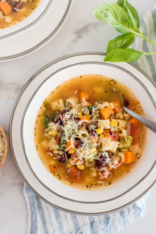 Overhead shot of Instant Pot minestrone soup in a white bowl, next to fresh basil and on a white and green striped cloth.