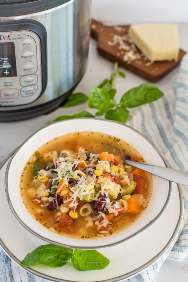 Overhead shot of Instant Pot Minestrone soup in a white bowl, with a basil garnish, parmesan, and an Instant Pot in the background.