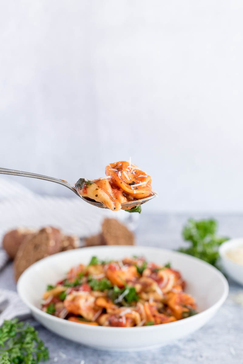 A spoonful of Instant Pot tortellini with fresh parmesan, with a white serving bowl filled with tortellini in the background.